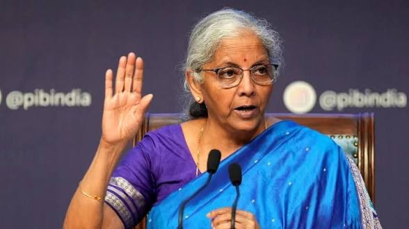 Finance Minister Nirmala Sitharaman speaks at a press conference, gesturing with her right hand, with the "@pibindia" social media handle visible in the background, as she discusses India's completed corporate bond tokenization pilot and urges the RBI to expand CBDC and digital rupee initiatives.