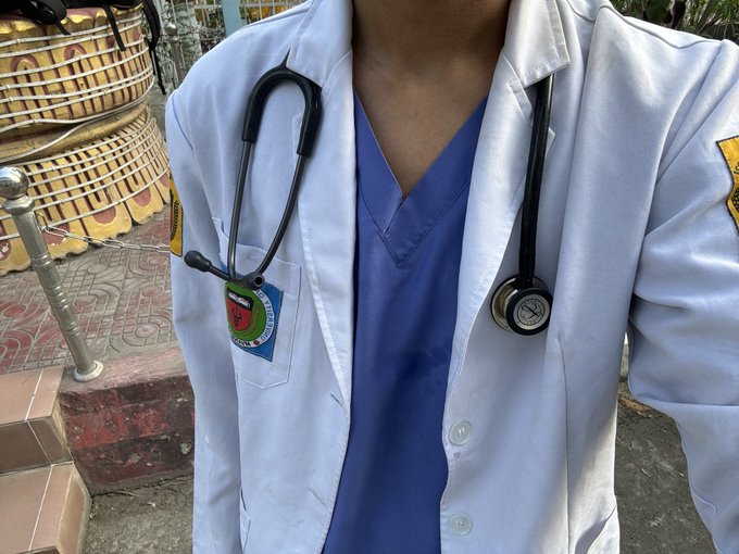 A person wearing a white medical lab coat with a stethoscope around their neck, blue scrubs underneath, and an ID badge with an Indonesian flag pin, photographed outdoors near what appears to be a decorative structure.