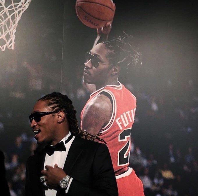 A man wearing sunglasses and a tuxedo stands in front of a large poster featuring a basketball player in a red 'FUTURE' #2 jersey about to dunk.