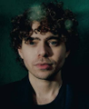 The image shows a man with curly hair looking directly at the camera, wearing a black shirt and standing against a dark background that resembles a green wall or green tent. The accompanying text seems to be related to a social media platform called Polymarket where people are paying $3k/month to post fake videos of themselves in exchange for money. This suggests that some individuals are making money by creating content on this platform.