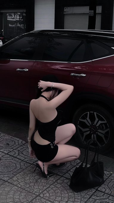 A woman in a black dress crouches beside a dark red SUV on a tiled street, with a black handbag beside her, embodying the post's themes of fleeting beauty and resilience amid struggle, tagged with @NomismaNetwork and @useTria.