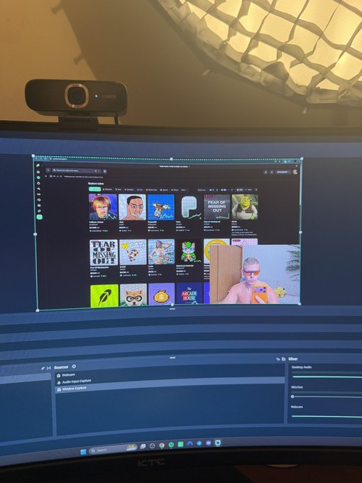 The image shows a curved gaming monitor displaying what appears to be a streaming or content creation interface (OBS), with a webcam mounted on top and artwork featuring "Fear of Missing Out" NFT-style images visible on screen. It relates to the caption about streaming "2 days in a row," suggesting the setup is used for live streaming content.