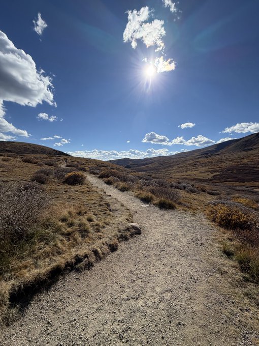 A scenic landscape photo shows a dirt trail winding through a grassy, rolling hillside under a bright sun and a vivid blue sky scattered with clouds.