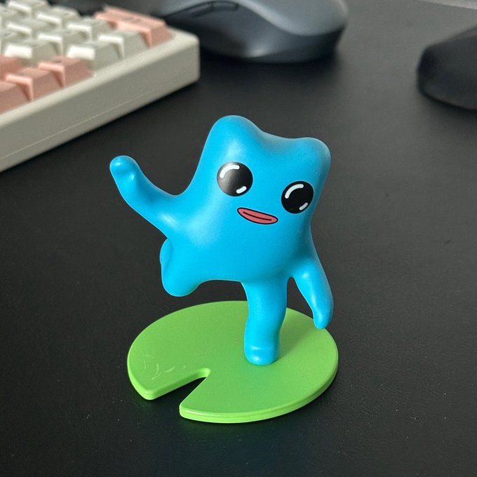 A small blue cartoon-style figurine with a smiling face stands on a green lily pad-shaped base, positioned on a dark desk in front of a keyboard and computer mouse.