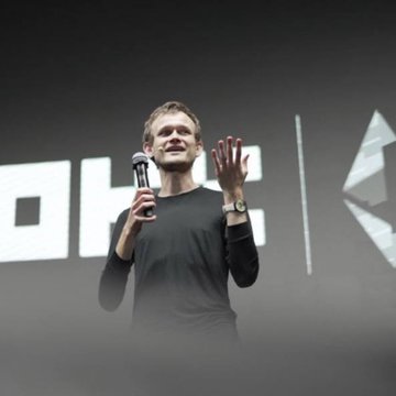 A man in a black long-sleeve shirt speaks on stage while holding a microphone, gesturing with his other hand, in front of a backdrop displaying the OKX logo. The image aligns with the post capturing the speaker at an OKX event in Singapore, where he shares his vision for the future of markets and money.