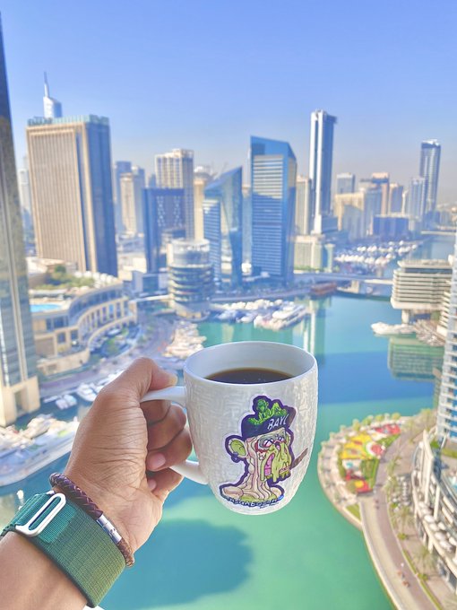 A hand holds up a sticker-covered coffee mug against a stunning aerial view of a modern marina city skyline, with a melting zombie character design reading "BARCELONA" as a playful nod to Monday morning resilience.