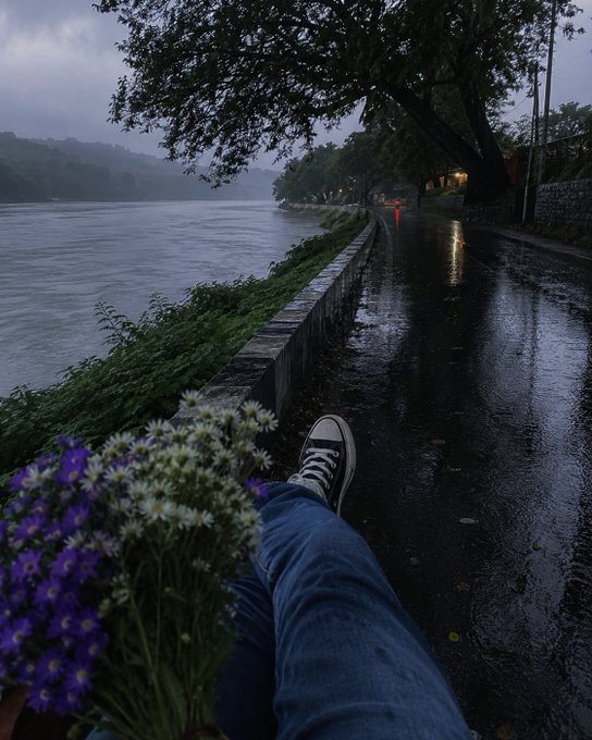 The image shows a person sitting on a bench with their legs crossed, holding flowers next to them. The scene takes place near a river or lake, possibly during a rainy day. There are also some purple flowers visible in the image. The photo is related to the post because it features an image of a person's feet and a bouquet of flowers, which could be interpreted as a metaphor for the connection between Web3 and users' daily lives.