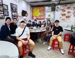 A group of about ten young people posing together indoors at what appears to be a restaurant, with framed pictures and a menu on the wall behind them.