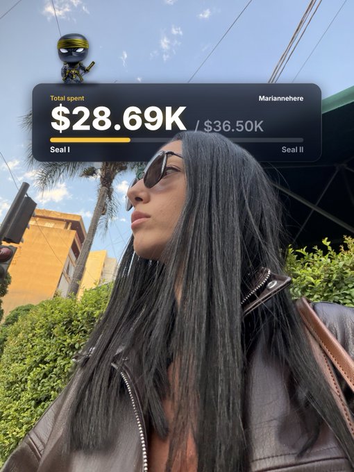 A selfie of a young woman with long dark hair wearing sunglasses and a leather jacket, with an overlay showing 'Total spent $28.69K / $36.50K' for user 'Mariannnehere' progressing toward 'Seal II'.