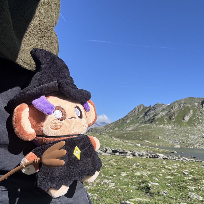 A plush monkey toy dressed as a witch with a black pointed hat and robe, holding a tiny broomstick, poses against a stunning alpine landscape with rocky mountains, a lake, and bright blue sky.

The image perfectly illustrates the "Magic Chimpi on a mountains adventure !CHIMP" theme, showcasing the whimsical character exploring a breathtaking high-altitude wilderness setting.