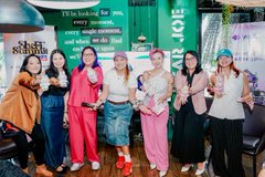 A group photo of seven women posing together at what appears to be a Thai cultural event or fair, with banners and signage in the background.