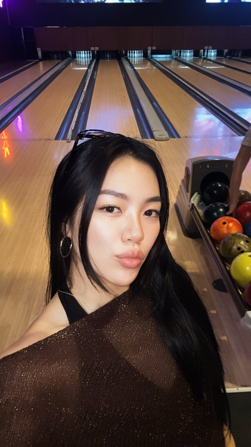 A selfie of a young woman with long dark hair wearing a brown sparkly top at a bowling alley, with bowling lanes and colorful balls visible in the background.
