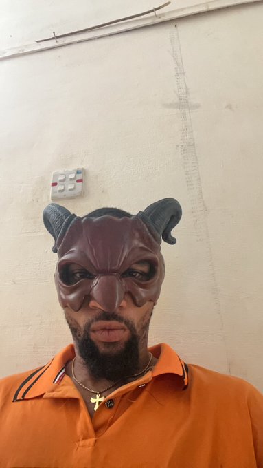 A man with a beard wearing an orange polo shirt and a red devil mask pushed up on his forehead stands against a plain wall with a light switch panel.