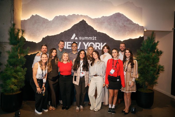 A group of about eleven people pose together smiling in front of a large backdrop featuring a mountain silhouette and the text "Summit NEW YORK," flanked by potted evergreen trees, capturing the Avalanche (AVAX) marketing team at the conference.