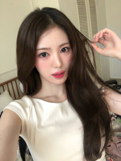 A selfie of a young woman with long brown hair and pink makeup, wearing a white t-shirt and playing with a strand of her hair.
