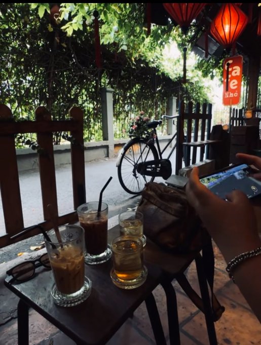 The image shows a person sitting at an outdoor table with three glasses of drinks, including two iced tea drinks and one coffee drink. The table also has a sandwich placed next to the drinks. A bicycle can be seen parked nearby, adding to the relaxed atmosphere of the scene.