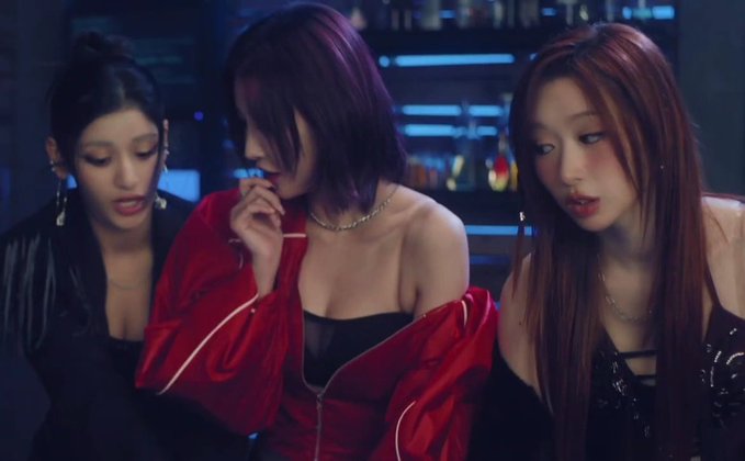 Three young women in edgy fashion, including one in a bright red off-the-shoulder jacket, are clustered together in a dark room with blue ambient lighting.