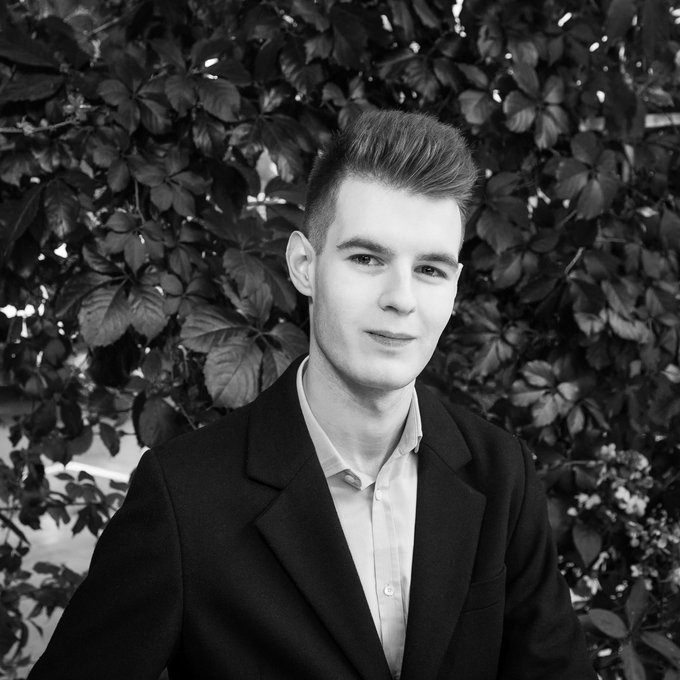 The image features a young man wearing a suit, standing against a backdrop of leaves. The photo has been edited with black and white tones, giving it an artistic touch. In the background, there are leaves that add depth and texture to the scene. The caption "Launch up only token = scammer Launch useless down only token= omg vitalik u the best" is present in the image, which seems unrelated to the main subject of the photo - the young man dressed in a suit.