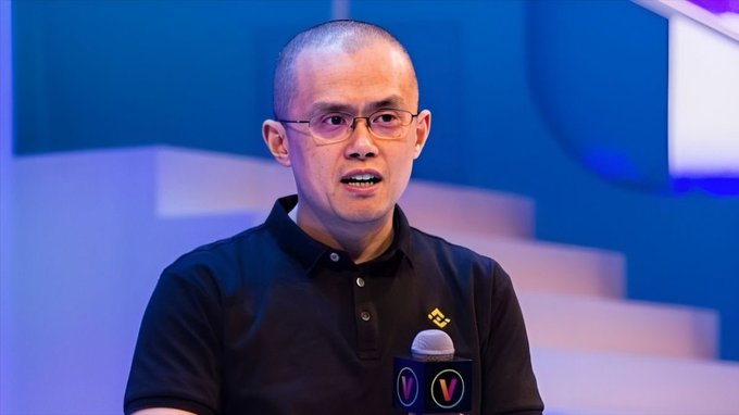 A man with glasses and a shaved head wearing a black polo shirt speaks at what appears to be a Binance event, with a microphone bearing a 'V' logo visible.