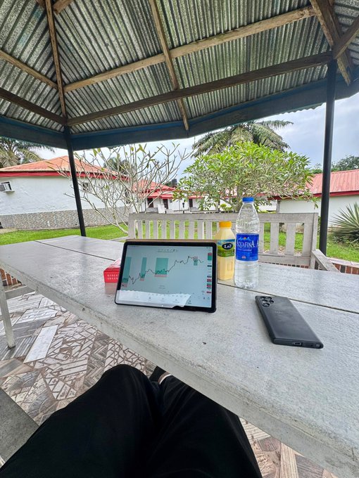 A tablet displaying a financial trading chart sits on an outdoor table alongside bottles of water and a phone, with lush greenery visible in the background.