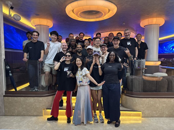 A group photo of approximately 20 people smiling and posing together in a futuristic, curved room with illuminated ceiling fixtures, blue screens, and many wearing black t-shirts with a white logo.