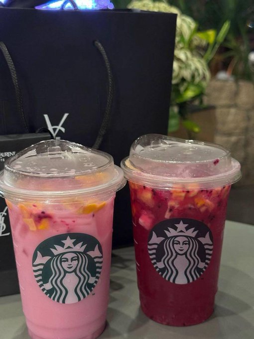 The image features two Starbucks drinks sitting side by side, each with a clear lid that allows for a view of their contents. The drinks are placed on a table, possibly at a café or similar establishment. The background includes some potted plants, adding a touch of greenery and life to the scene.