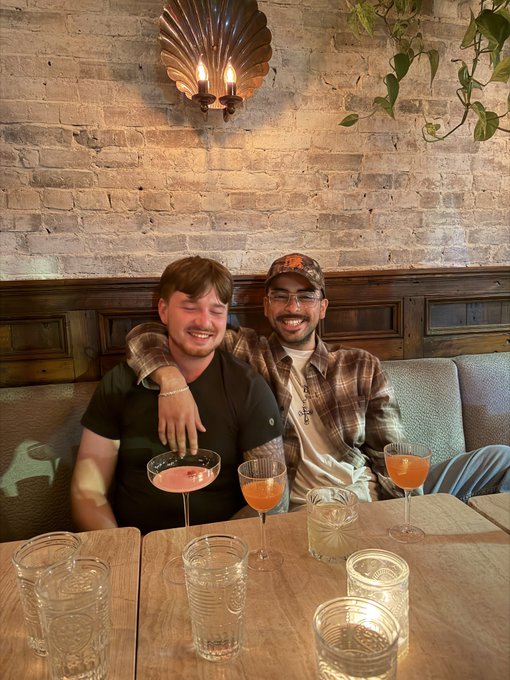 Two smiling men sit closely together at a table in a restaurant booth with pink cocktails and water glasses in front of them.