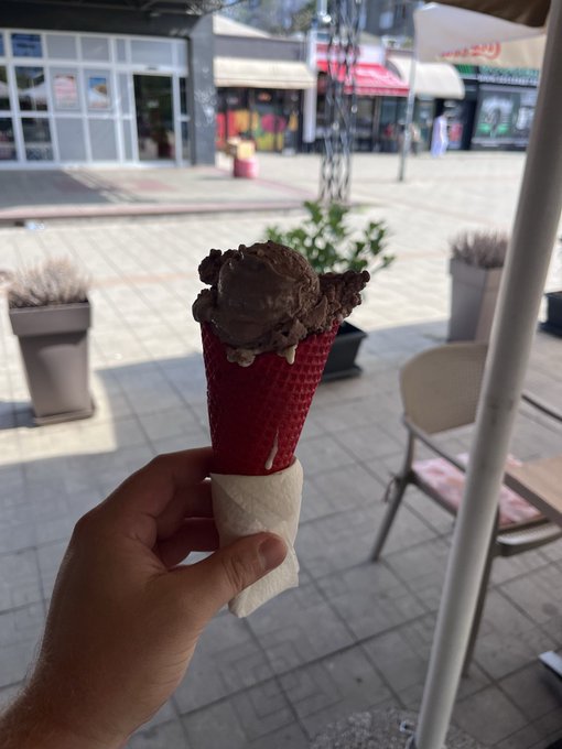 A hand holds a red waffle cone filled with chocolate ice cream wrapped in a napkin, set against an outdoor plaza with storefronts in the background.