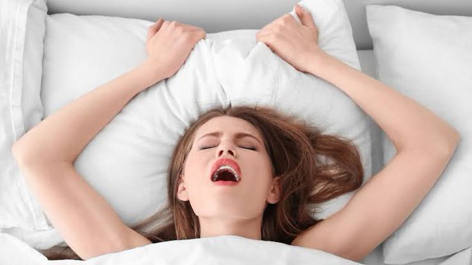 A young woman lying in bed with her arms stretched up, yawning widely while surrounded by white pillows and blankets.