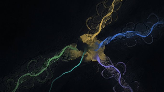 An abstract artistic image featuring flowing, smoke-like streams of green, yellow, blue, and purple emanating from a central point against a dark background.