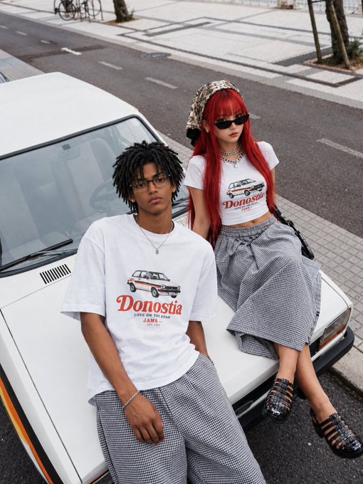 Two young people in streetwear posing on a white car, wearing matching 'Donostia' graphic t-shirts featuring a van design, paired with checkered bottoms.