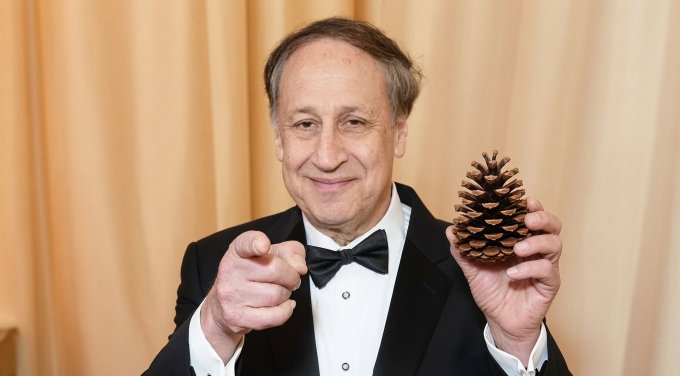 A smiling older man in a tuxedo points at the camera with one hand while holding up a large pinecone in the other, set against a beige curtain backdrop. The image humorously ties to the AMC "popcorn ape" post by visually representing the pinecone as a punchline, riffing on the meme of Adam Aron teasing retail investors with more turbulence ahead.