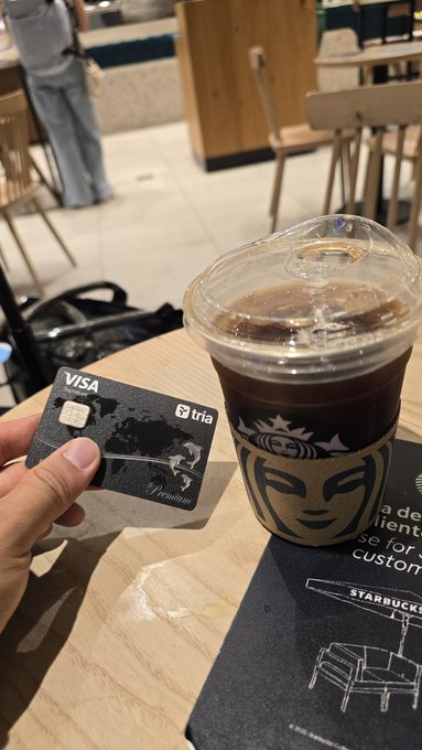 A hand holds a black Visa credit card from 'tria' next to a Starbucks iced coffee cup on a table inside a café, with a Starbucks mat visible underneath.