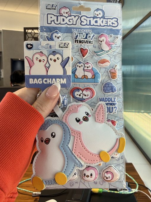 A hand holding 'Pudgy Penguins' merchandise including a 'BAG CHARM' and 'PUDGY STICKERS' featuring cute penguin character designs in pastel colors.