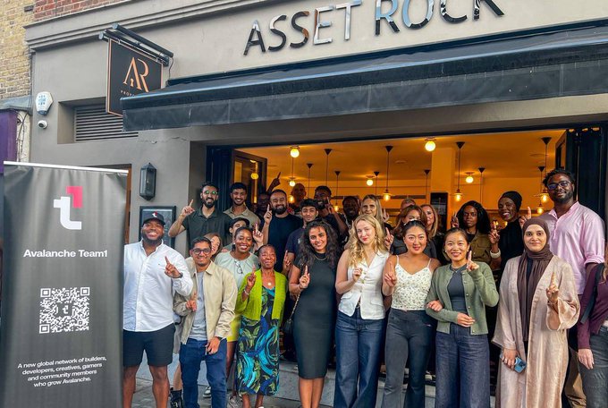 A large group of people poses outside a venue called 'ASSET ROCK,' standing next to a banner for the 'Avalanche Team' featuring a QR code and describing it as a network for builders, developers, and creators.