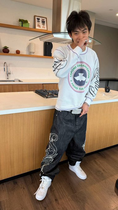 A young man stands in a kitchen wearing a white long-sleeve shirt with a circular logo reading "MORONS ORGANIZATION" and a star, dark jeans with decorative stitching, white sneakers, and a sparkly belt.
