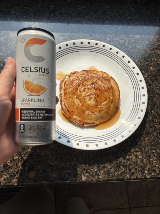 A hand holds a can of Celsius Sparkling Orange energy drink next to a plate of swirled pancakes drizzled with syrup on a granite countertop.