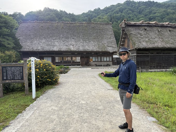 A man wearing a navy blue jacket, gray shorts, and sunglasses stands on a path in front of traditional Japanese thatched-roof houses with forested hills in the background.