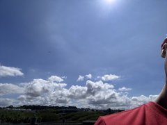 Blurry selfie showing a person's arm in a red shirt against a blue sky with white clouds over a landscape.
