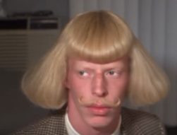 A person with an exaggerated blonde bowl-cut wig and curled mustache stares blankly forward, humorously representing a trader looking unimpressed or bewildered while reviewing a market chart.