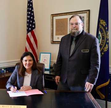 Photo of a smiling woman seated at a desk signing a document, with a bearded man in a suit standing beside her in an official office setting with American and state flags.