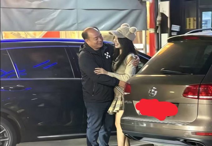 An older man in a dark jacket embraces a young woman wearing a bear-eared beanie and plaid skirt, standing between two parked SUVs at night. The scene appears to capture a candid street encounter, which the accompanying post suggests may be a photo of someone impersonating crypto figure "Xu Mingxing" (徐明星) to flirt with the woman.