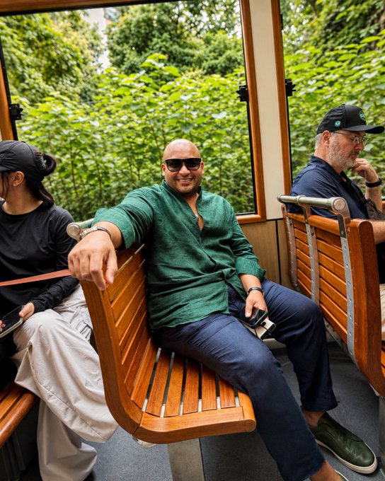 A man in a green shirt and sunglasses relaxes on a wooden cable car seat, smiling at the camera as lush greenery passes by outside, while two other passengers sit nearby enjoying the ride through Hong Kong's scenic trails. The casual travel vibe captures the relaxed yet optimistic spirit of attending Bitcoin Asia 2026, with his smile reflecting the post's bullish outlook on the conference experience.