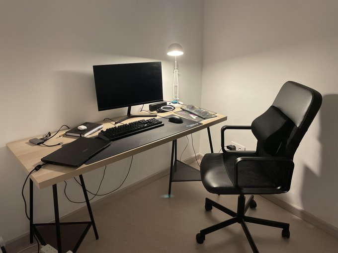 A minimalist home office setup features a wooden desk with a monitor, keyboard, and lamp, paired with a black office chair in a corner room.