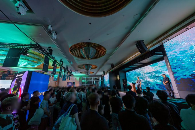 A crowded indoor event space features people gathered for a presentation, with a large screen on the right displaying an underwater scene and the partial text 'Immire to' and 'Kai Finance.'
