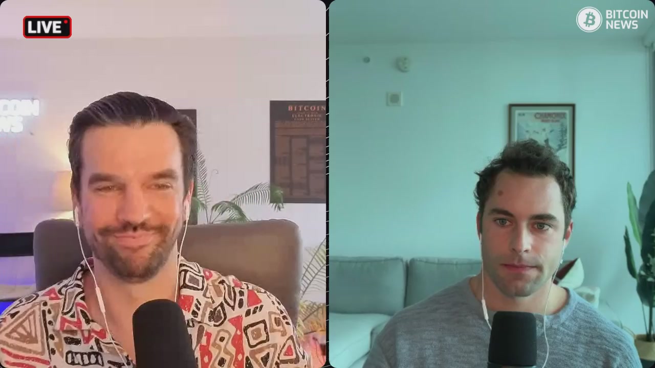 The image shows two men sitting behind microphones, each holding a microphone with their hands placed over them. They appear to be engaged in a conversation or presentation, possibly discussing topics related to Bitcoin or other financial matters. The background features a white wall with a poster on it and a potted plant nearby.
