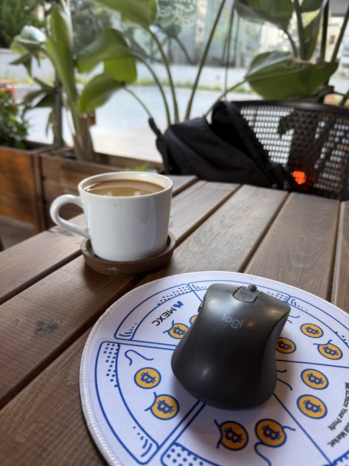 A cup of coffee sits on a wooden table beside a Logitech mouse resting on a colorful MEXC-branded mousepad, capturing a quiet work-from-café moment during an unexpected layover.