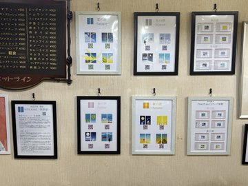 A display of framed collectible stamp sheets and certificates mounted on a wall, alongside a dark wooden sign with Japanese text and pricing information for what appears to be a stamp collection or philatelic display.