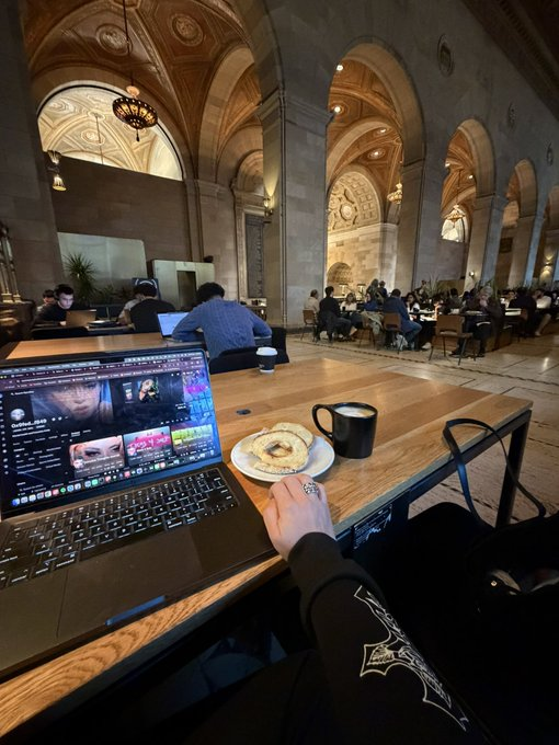 The image shows a person sitting at a table with an open laptop computer, eating breakfast while working or browsing the internet. There are other people present in the room as well, but they do not seem to be interacting with each other. The scene appears to be taking place inside a building that has multiple arches and columns, giving it a grand appearance. The person's attire consists of a black shirt paired with a white baseball cap.