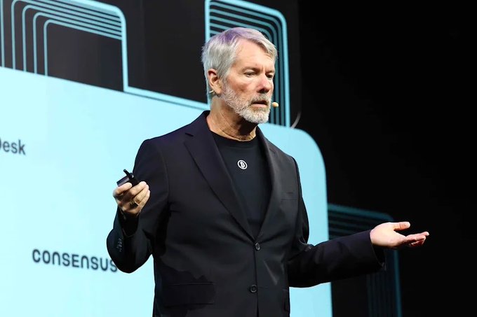 A gray-haired man in a black blazer speaks at a 'CONSENSUS' conference, gesturing with his palm open while holding a presentation remote.